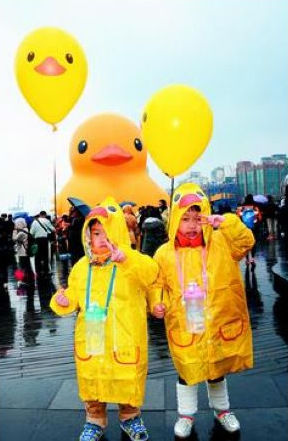 两名小朋友在小鸭前面拍照，全身配备小鸭各种装备，帽子、气球、水壶等一应俱全